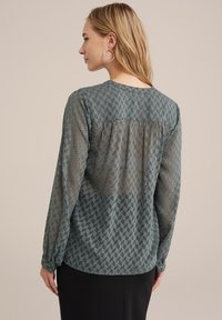 Bluză verde transparentă cu un model geometric, mâneci lungi și manșete cu butoni. Decolteu rotund cu detalii strânse pe spate.