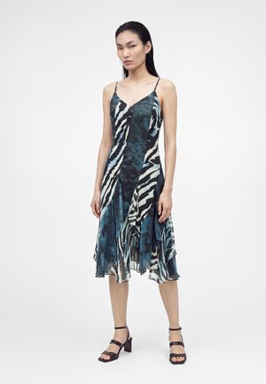 Femme portant une robe asymétrique sans manches avec un imprimé zèbre bleu et noir, debout avec des sandales noires à talons blocs ouvertes à l’avant sur fond blanc.