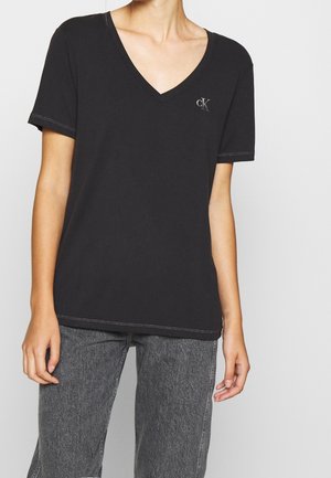 Person wearing a black V-neck short-sleeve T-shirt with small "ck" logo on left chest and gray high-waisted jeans.