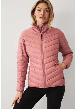 Vrouw draagt een roze doorgestikte jas met ritssluiting en opstaande kraag, met één hand in de zak en zwarte broek, tegen een effen lichte achtergrond.