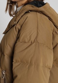 Femme portant une doudoune d'hiver beige avec une capuche noire et une fermeture éclair en métal visible sur la manche.