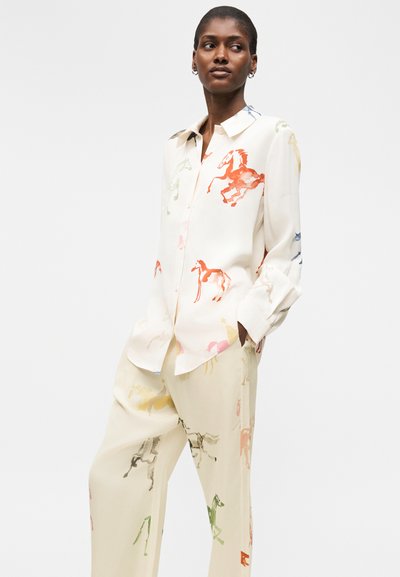 Γυναίκα που φοράει άσπρο φαρδύ πουκάμισο και παντελόνι με πολύχρωμα σχέδια αλόγων, στέκεται με τα χέρια στις τσέπες και κοιτάζει προς το πλάι.