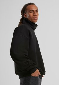 Svart fleecegenser med glidelås og høy krage, avslappet passform og ribbestrikkede mansjetter. Myk tekstur og minimalisk design, egnet for uformell bruk.