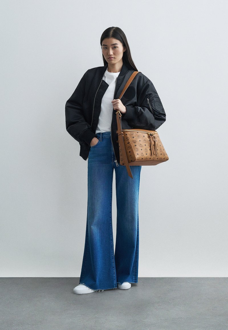 Frau in schwarzer Bomberjacke, weißem Shirt, weitgeschnittenen blauen Jeans, weißen Sneakern, die eine sandfarbene Designertasche mit Schultergurt hält.