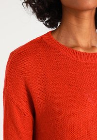 Červený pletený svetr s žebrovaným kulatým výstřihem, měkkou texturou a volným střihem, s viditelnými švy a mírným leskem.