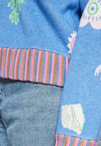 Blå strikket sweater med blomster mønstre og en flerfarvet stribet ribkant. Stoffet virker blødt og tekstureret.