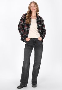 Veste à carreaux noire et rouge avec des accents gris, portée sur un haut beige, associée à un jean gris foncé et des chaussures noires. Tissu lisse avec une coupe structurée.