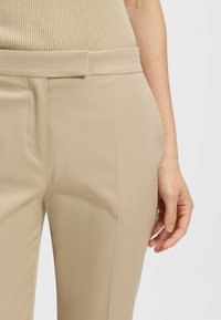 Beige maßgeschneiderte Hose mit glatter Textur, schlankem Schnitt und seitlichen Taschen. Der Bund ist flach und nahtlos konstruktiert für klare Linien.