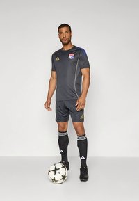 Mies jalkapalloilija mustassa varustuksessa, jossa on Lyonin logo, seisoo yksi jalka valkoisen ja mustan jalkapallon päällä tasaisella taustalla.