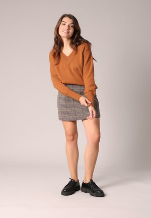 Un pull couleur rouille avec un décolleté en V, associé à une jupe mini à carreaux. Des chaussures noires à lacets complètent la tenue. Fond simple.