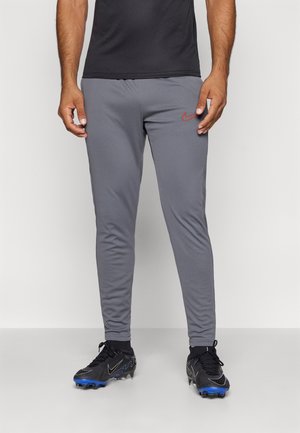 Mann trägt graue Slim-Fit Nike-Sporthose und schwarze Fußballschuhe mit blauen Stollen, steht vor einem einfarbigen weißen Hintergrund.