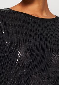 Černý síťovaný top se lesklými flitry, s pohodlným výstřihem a krátkými rukávy. Látka má texturovaný, vzorovaný vzhled.