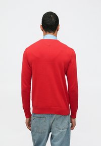 Άνδρας φορώντας κόκκινο πουλόβερ με μακριά μανίκια πάνω από μπλε πουκάμισο και ανοιχτό μπλε τζιν, στέκεται απέναντι σε απλό λευκό φόντο.