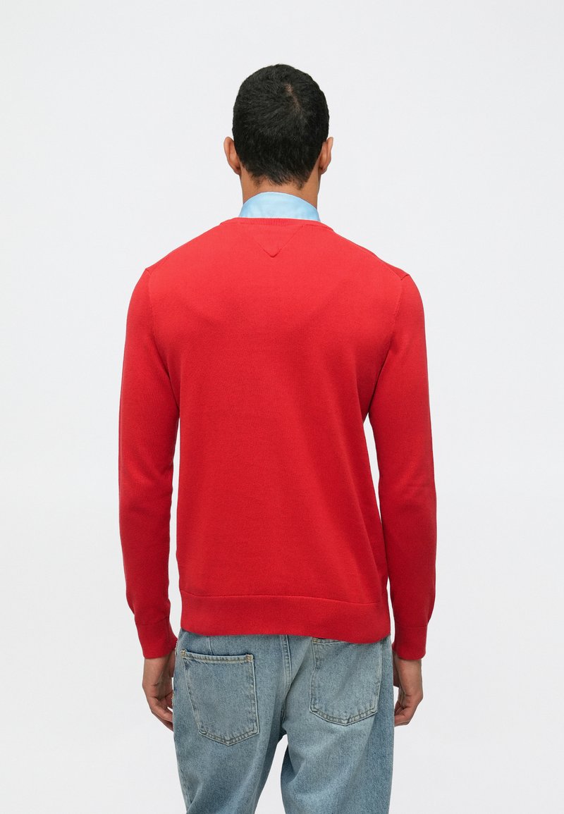 Άνδρας φορώντας κόκκινο πουλόβερ με μακριά μανίκια πάνω από μπλε πουκάμισο και ανοιχτό μπλε τζιν, στέκεται απέναντι σε απλό λευκό φόντο.