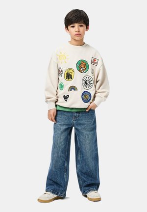 Crème-kleurige sweatshirt met verschillende kleurrijke patches, gecombineerd met blauwe denim wijde jeans en witte sneakers. Groene shirt zichtbaar eronder.