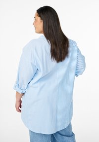 Camicia oversized a righe blu con maniche arrotolate, caratterizzata da righe verticali e orizzontali, tessuto morbido e orlo curvo.