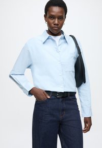 Svetlomodrá košeľa na zapínanie s golierom, dlhými rukávmi a náprsným vreckom, v kombinácii s tmavomodrými džínsami. Súčasťou je čierny opasok a kabelka.