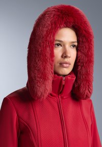 Piros dzseki texturált mintával, puha, szőrmével bélelt kapucnival. Az anyag simának tűnik, és a cipzár középen található.