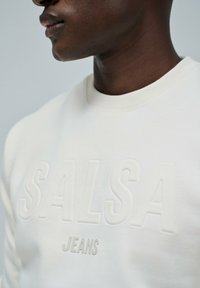 Close-up van een persoon die een wit sweatshirt draagt met het reliëf "SALSA" en verhoogde "JEANS" logo's op de borst.
