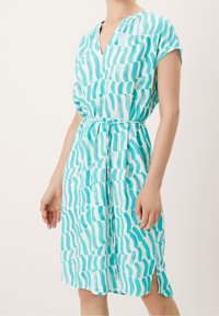 Une robe midi turquoise à motif ondulé, avec des manches courtes, un col en V, une ceinture à nouer à la taille, et une fente latérale pour plus de confort.