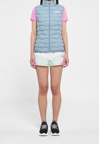 Gilet imbottito azzurro chiaro con colletto alto, cerniera frontale, indossato sopra una t-shirt rosa e pantaloncini multicolore. Scarpe da ginnastica nere con suole bianche.