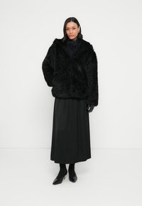 Czarna kurtka z czarnego futra ekologicznego o pluszowej fakturze i szerokim kołnierzu, zestawiona z długą czarną spódnicą i czarnymi botkami za kostkę.