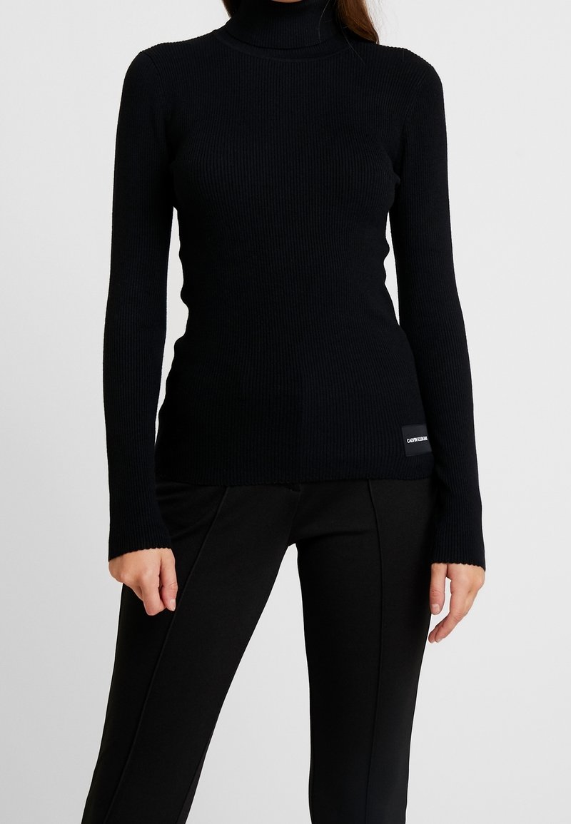 Pull noir côtelé à col roulé avec manches longues, coupe ajustée et petit patch logo discret en bas à gauche. Texture lisse.