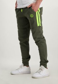 Olijfgroene joggingbroek met twee verticale neongele strepen, voorzakken, logo detail en een taps toelopend ontwerp. Gekenmerkt door het dragen van witte sneakers.