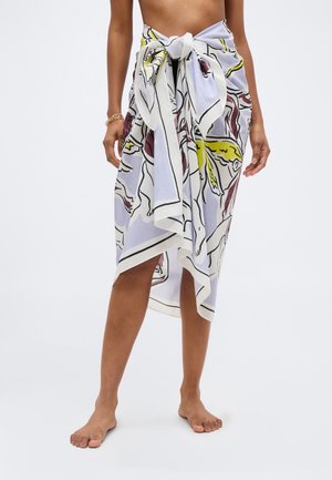 Femme portant un paréo à imprimé abstrait pastel noué à la taille, pieds nus, vue de la moitié du torse aux pieds sur fond blanc.