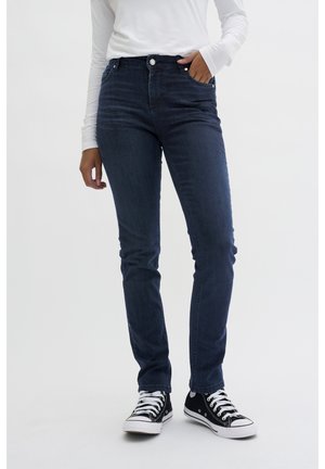 Jeans in denim blu scuro con vestibilità slim, vita media, design a cinque tasche e una texture liscia, abbinati a sneakers nere.
