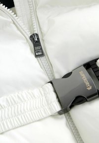 Balta pūkaina jaka ar pelēku sprādzi un rāvējslēdzēju, uz kura ir "SUPERDRY" zīmols. Gluda tekstūra ar savilktiem elastīgiem akcentiem.