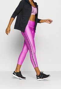 Idrottskläder som består av en rosa sport-bh, glänsande rosa leggings med svarta sidostreck och en svart zip-up hoodie. Svarta sneakers fulländar looken.