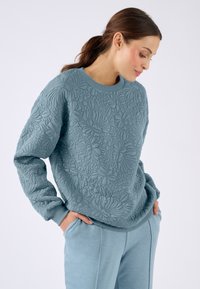 Mujer con pelo oscuro en una coleta, vistiendo un sudadera acolchada azul con estampado floral y pantalones azules a juego, con las manos en los bolsillos, mirando hacia abajo.