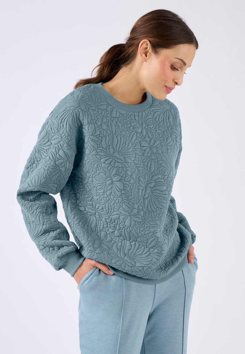 Mujer con pelo oscuro en una coleta, vistiendo un sudadera acolchada azul con estampado floral y pantalones azules a juego, con las manos en los bolsillos, mirando hacia abajo.