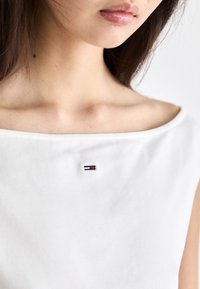 Valge puuvillane topp laia kaeluse ja lühikeste varrukatega, millel on rinnal väike punase, sinise ja valge värviga brodeeritud logo.