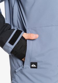 Wasserabweisende Jacke mit einem hellblauen Körper, schwarzen Ärmeln und einem Klettverschluss an den Manschetten. Enthält ein Logo-Patch und eine Seitentasche.