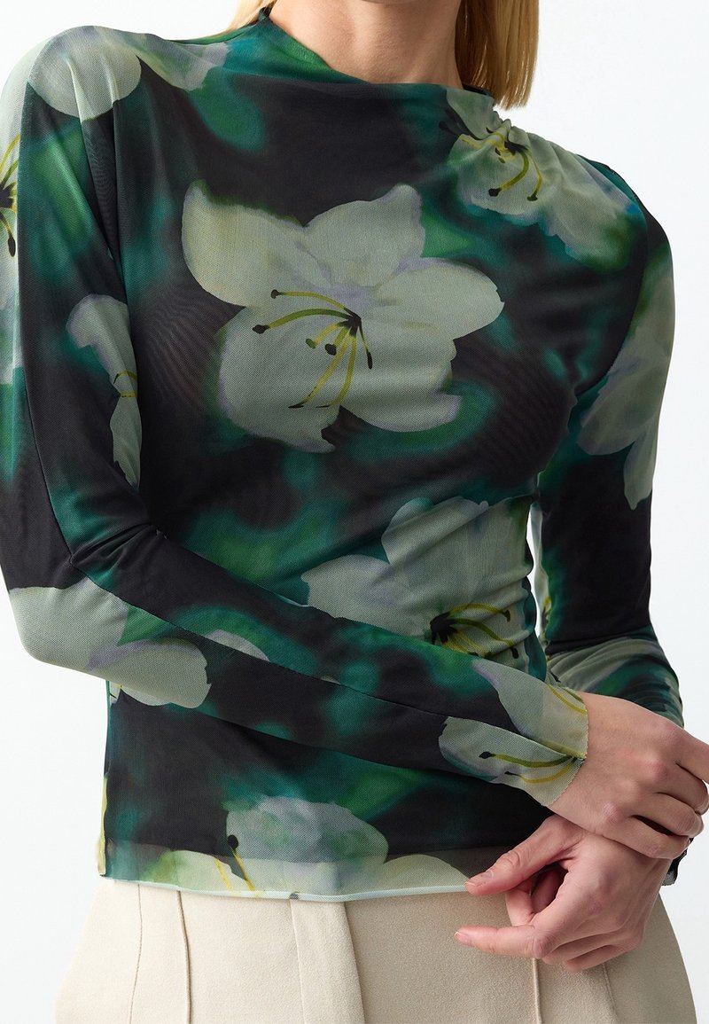 Femme portant un haut transparent à manches longues avec un grand imprimé fleur de lys blanche sur fond noir et vert, associé à un pantalon beige clair.
