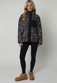 Kurtka polarowa w panterkę z wysokim kołnierzem, czarnym zamkiem błyskawicznym i kieszenią; noszona z czarnymi legginsami i brązowymi botkami za kostkę.