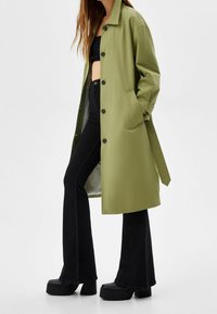 Olivgrön lång kappa med knappar, sidofickor och bälte. Bärs över en svart croppad topp och utsvängda svarta jeans, komplett med plattformsskor i svart.