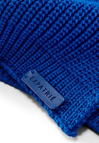 Gorro de punto azul con textura acanalada, hecho de hilo y con una etiqueta de cuero grabada con "EXPATRIÉ" pegada en el lateral.