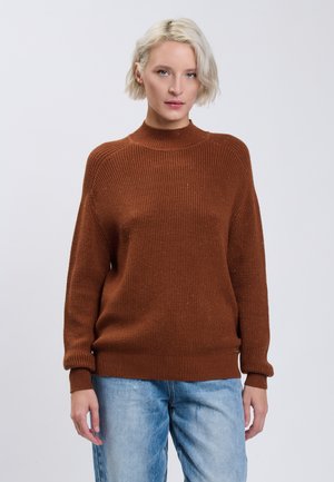 Giovane donna con capelli biondi corti che indossa un maglione marrone a coste e jeans azzurri, in piedi davanti a uno sfondo semplice.