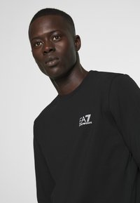 T-shirt nera a maniche lunghe in cotone, con un piccolo logo EA7 bianco sul lato sinistro del petto. Design semplice e classico con scollatura rotonda.