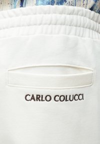 Biały materiał o fakturowanej powierzchni, z poziomą kieszenią i wyhaftowanym napisem "CARLO COLUCCI" w kolorze brązowym. Gładki i miękki wygląd.