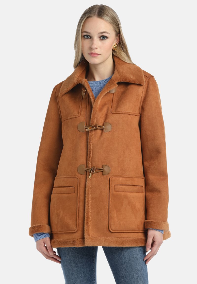 Cappotto in pelliccia sintetica marrone con ampio colletto, chiusure con fibbia, due tasche frontali e una consistenza morbida. Indossato su un maglione blu e jeans.