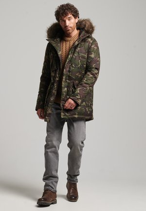 Tarnfarbenes Parka mit fellgefütterter Kapuze, Reißverschluss und Taschen. Getragen über einem Grobstrickpullover, kombiniert mit grauen Jeans und braunen Stiefeln.