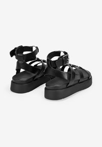 Sandalias de plataforma negras con múltiples correas cruzadas, hebilla ajustable en el tobillo, material de cuero suave y suela de goma texturizada.