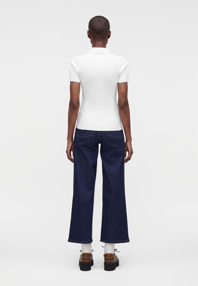 Personne aux cheveux courts portant un haut blanc à manches courtes, un jean large foncé, des chaussettes blanches et des chaussures plateformes marron, debout de dos.