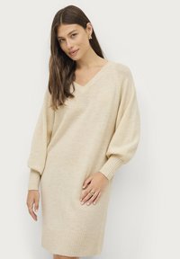 Beige gebreide trui jurk met een V-hals, losse mouwen en geribbelde zoom. Zachte textuur en tot halverwege de dij lengte. Minimalistisch ontwerp.