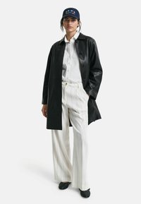 Schwarzer Ledermantel über einem weißen Hemd mit Knöpfen, kombiniert mit weiß gestreiften Hosen und einer dunklen Baseballmütze. Strukturierte schwarze Schuhe runden den Look ab.