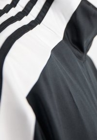 Svart och vit atletisk topp med hög krage, med tre vertikala vita ränder på axlarna och texturerade tygdetaljer.